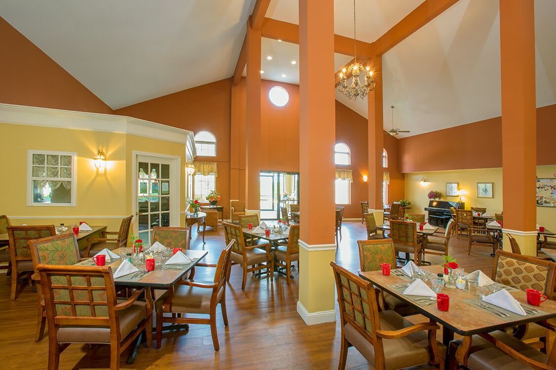 A spacious dining room with wooden floors and high ceilings, featuring elegantly set tables and chairs under warm lighting.