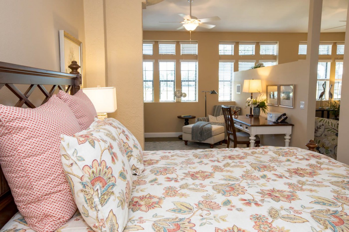 A cozy bedroom featuring floral bedding and soft lighting, with a view of a sitting area and large windows.