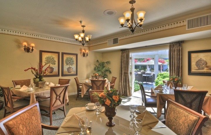 An elegantly furnished dining room with chandeliers and neatly set tables, overlooking a garden through large windows.
