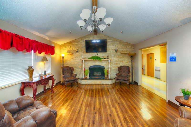 A cozy living room with hardwood floors, a fireplace, and a television mounted above. The space is furnished with armchairs and illuminated by a decorative chandelier.