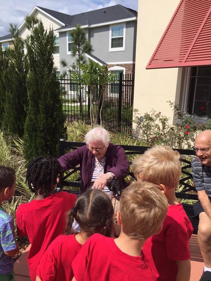 An elderly woman interacts with a group of children outside on a sunny day.