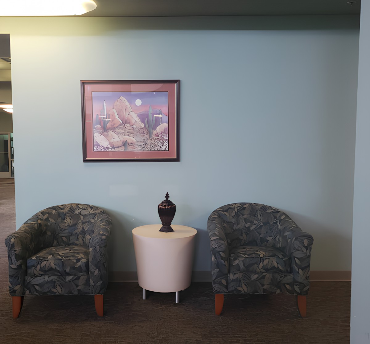 A cozy lobby area with two patterned chairs flanking a small round table topped with a decorative vase, and a framed artwork on the wall.