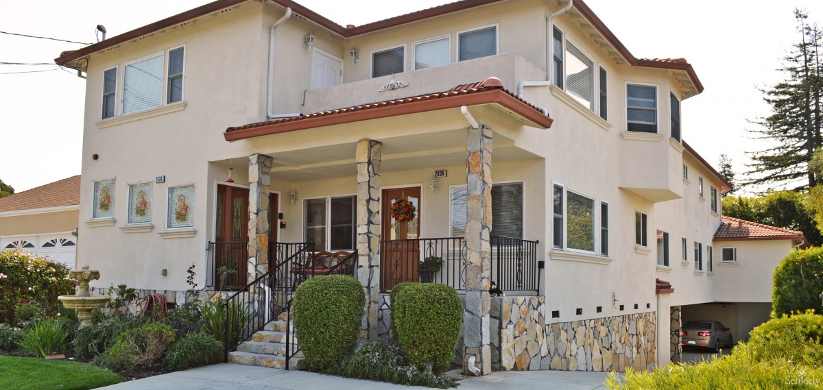 A two-story residential house with a front porch featuring stone columns and manicured shrubs.