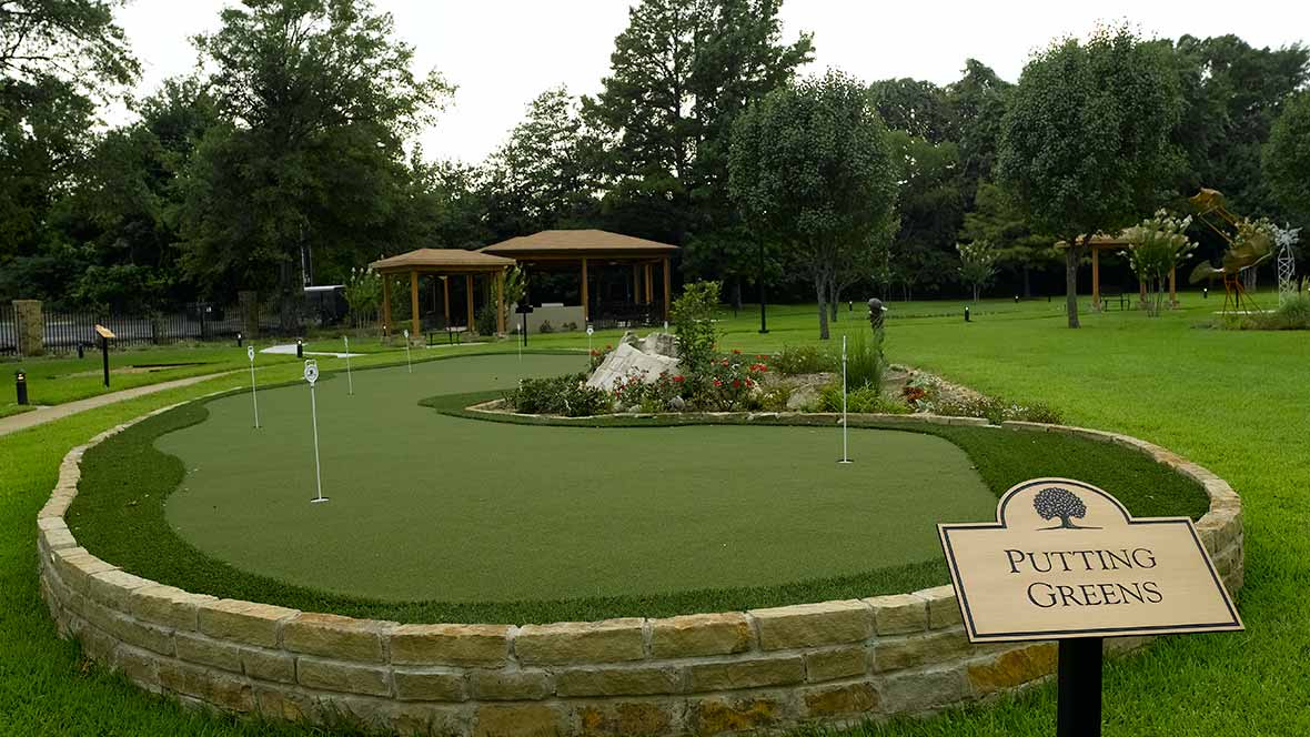 A beautifully landscaped outdoor putting green with surrounding greenery and two gazebos in the background.