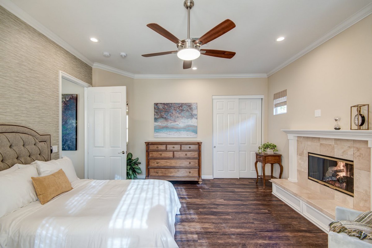 A cozy bedroom with wooden flooring, featuring a bed, dresser, ceiling fan, and fireplace.