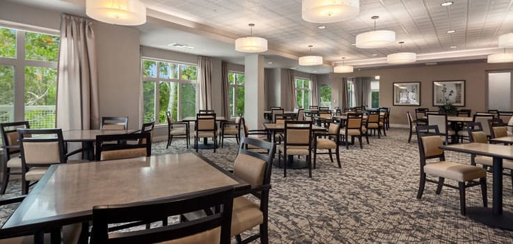 A well-lit dining area with neatly arranged tables and chairs, featuring large windows and modern lighting.