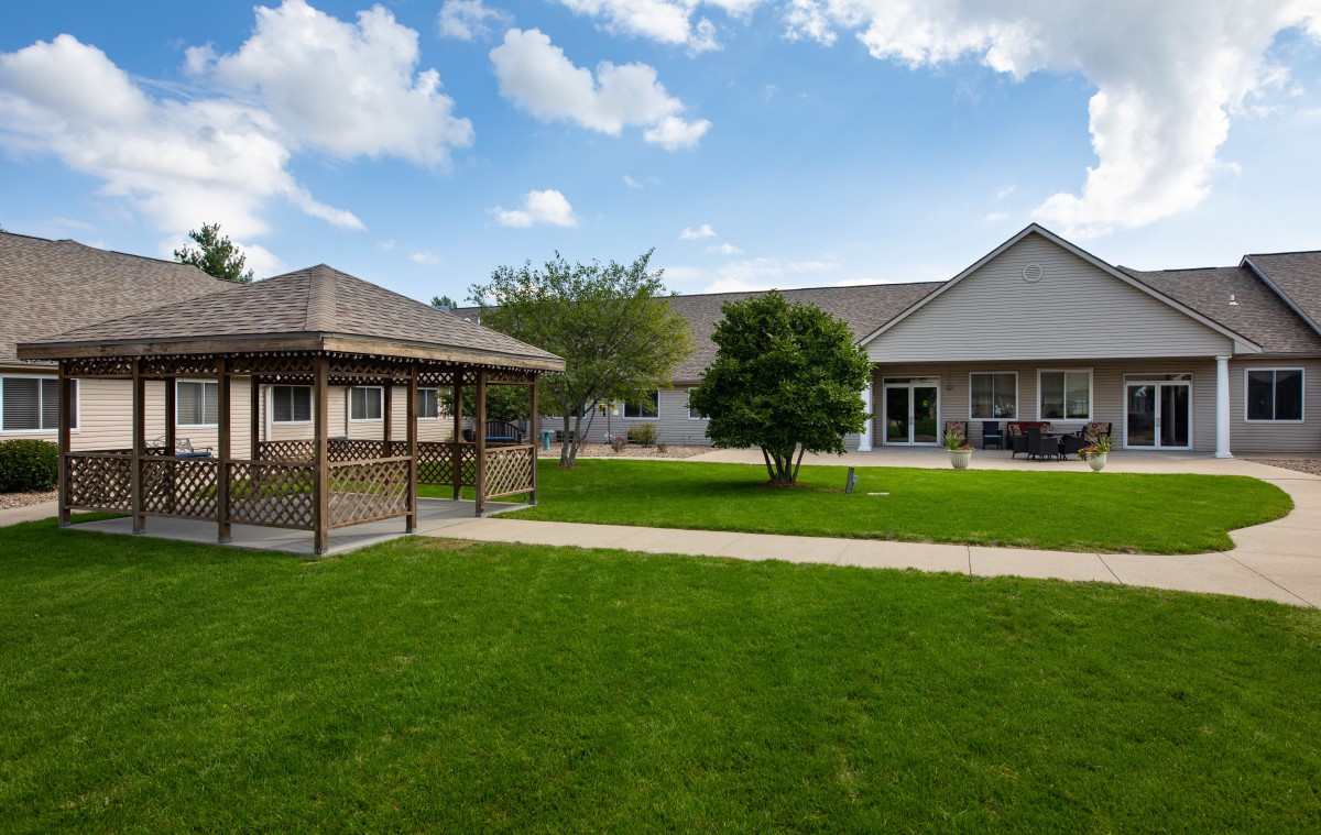 A peaceful backyard with a gazebo, green lawn, and a house under a blue sky with clouds.