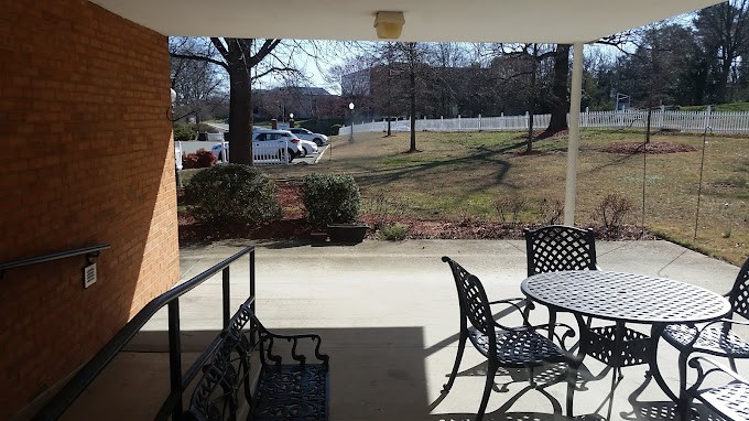 A view from a patio with metal chairs and a table, overlooking a grassy area with trees and a parking lot in the background.