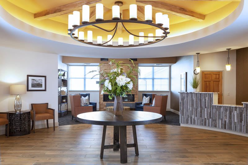 A modern lobby interior featuring a large chandelier, central table with flowers, and comfortable seating area.