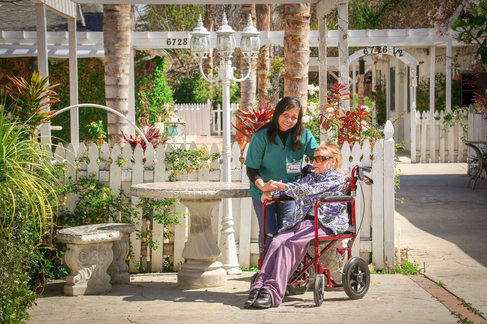 A caregiver assists an elderly person in a wheelchair in a garden setting.