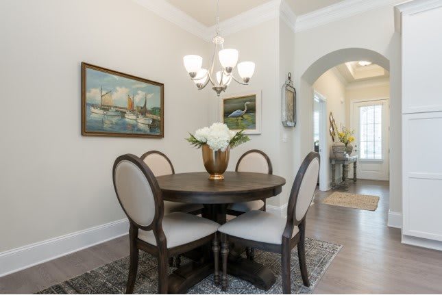 A cozy dining area featuring a round wooden table with four upholstered chairs, illuminated by an elegant chandelier. The walls are decorated with artwork, and a vase of flowers adds a touch of freshness to the setting.