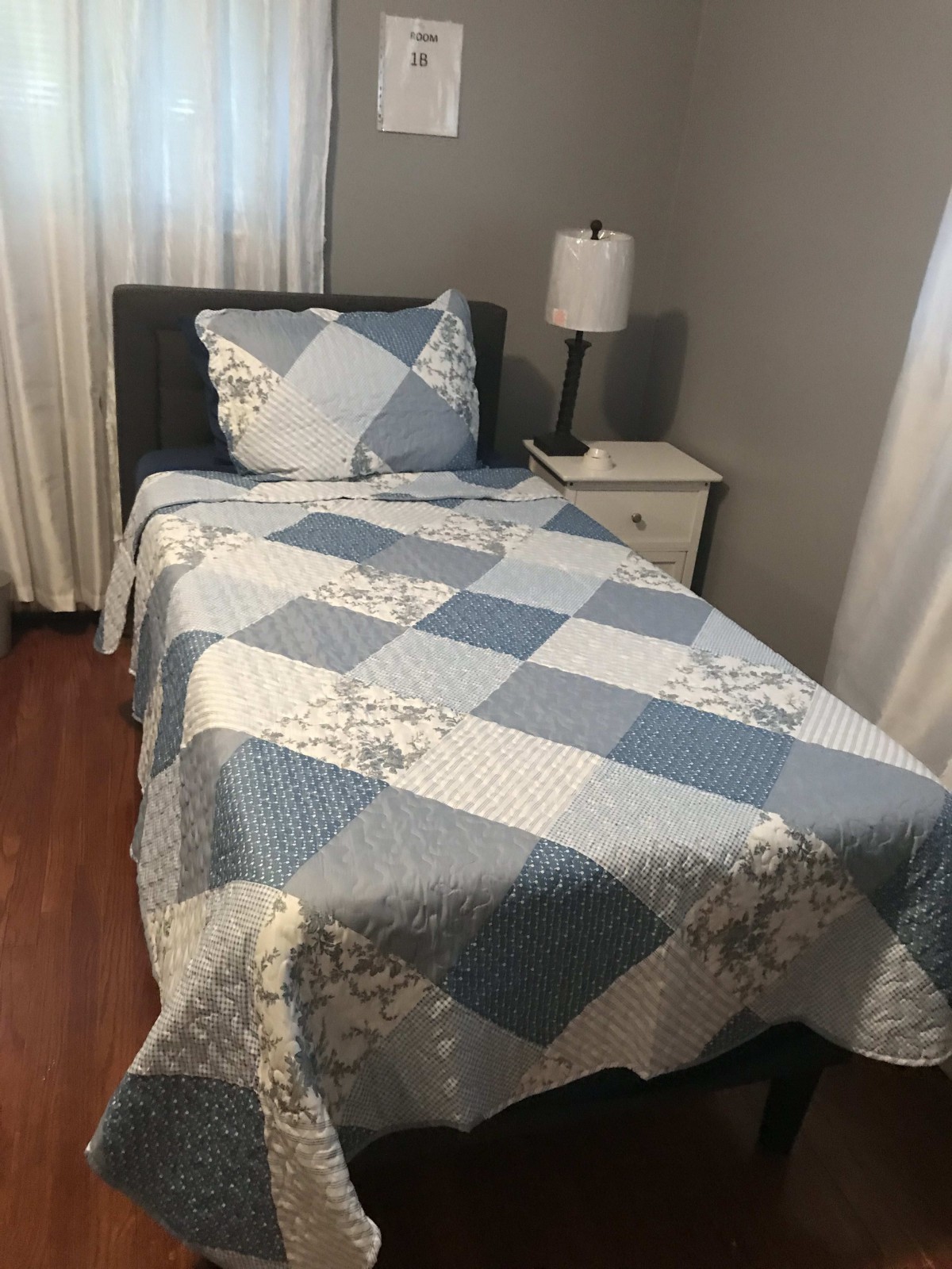 A neatly made single bed with a quilted cover, next to a nightstand and lamp in a cozy bedroom.