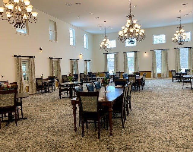 A spacious dining room with multiple tables and chairs, illuminated by elegant chandeliers and natural light from large windows.
