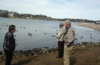 Two men fishing by a lake with ducks swimming nearby.