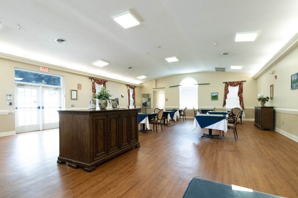 A spacious dining room with wooden flooring, featuring tables and chairs neatly arranged under bright lighting.