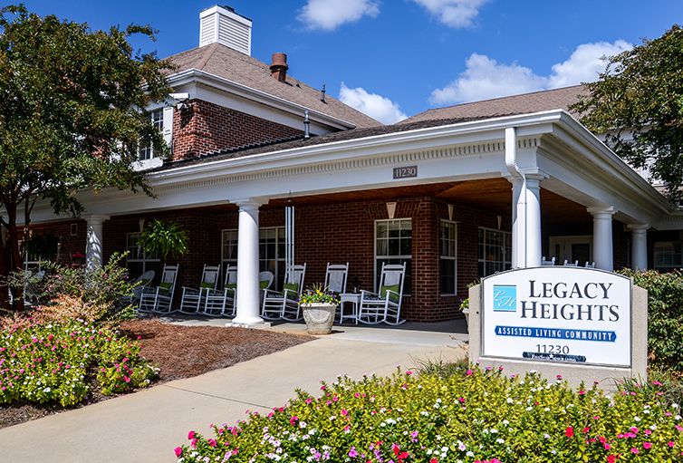 The entrance of a building with a porch featuring rocking chairs and a sign, surrounded by landscaped greenery under a blue sky.