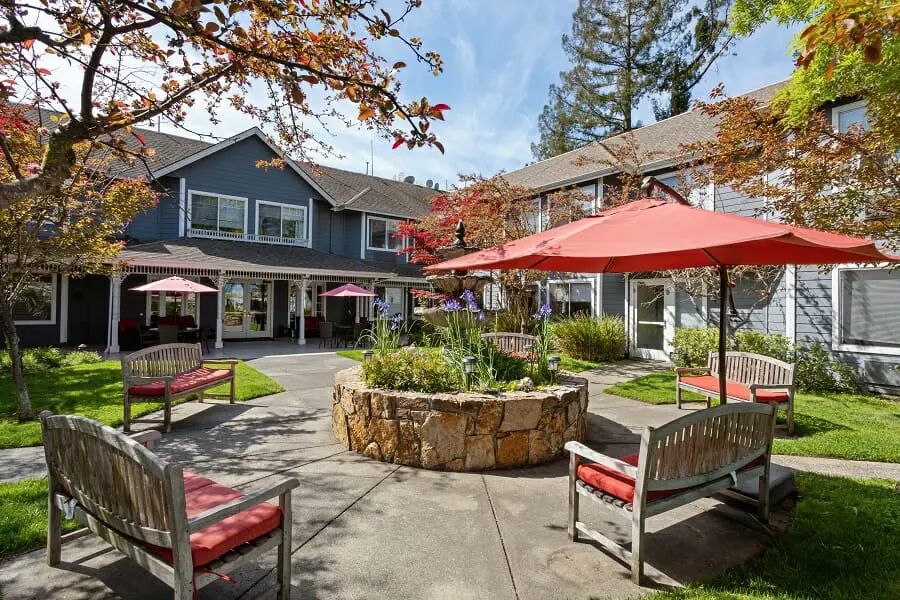 A peaceful courtyard featuring benches and umbrellas surrounded by a garden and building.