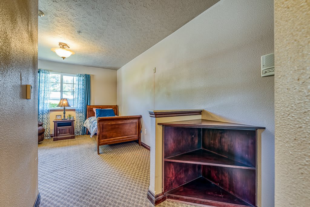 A cozy bedroom with a wooden bed frame, carpeted floor, and a corner shelf. The room is well-lit by a lamp and natural light from the window.