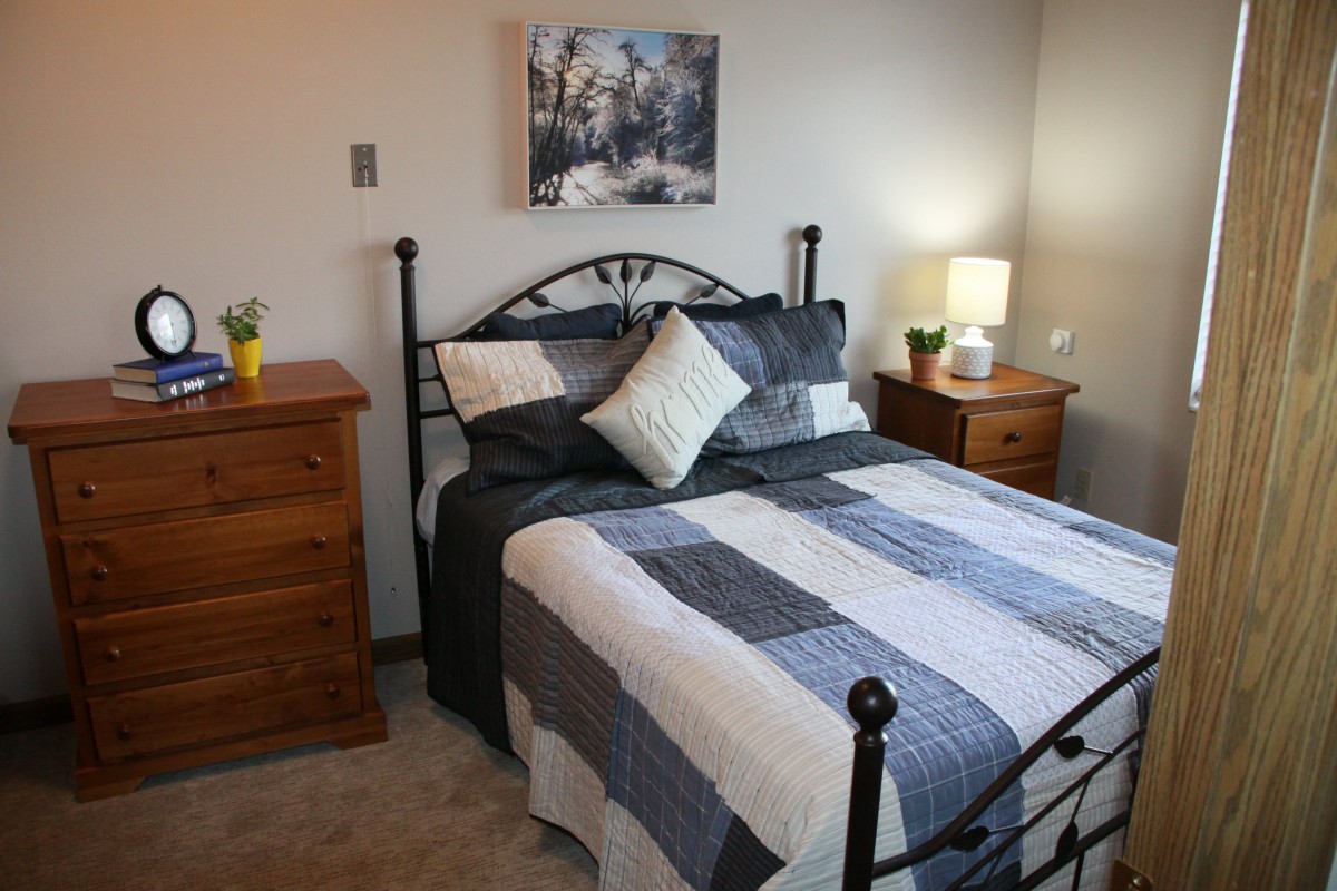 A cozy bedroom with a neatly made bed, flanked by wooden nightstands, each topped with a lamp and other decor. A winter-themed painting hangs on the wall above the bed.