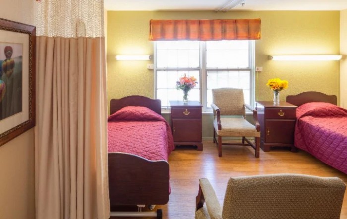 A brightly lit hospital room with a single bed, chairs, and floral decorations.