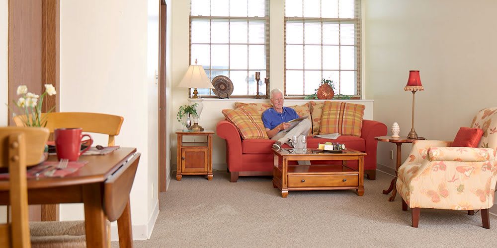 A cozy living room with a person sitting on a sofa, surrounded by warm decor and large windows.