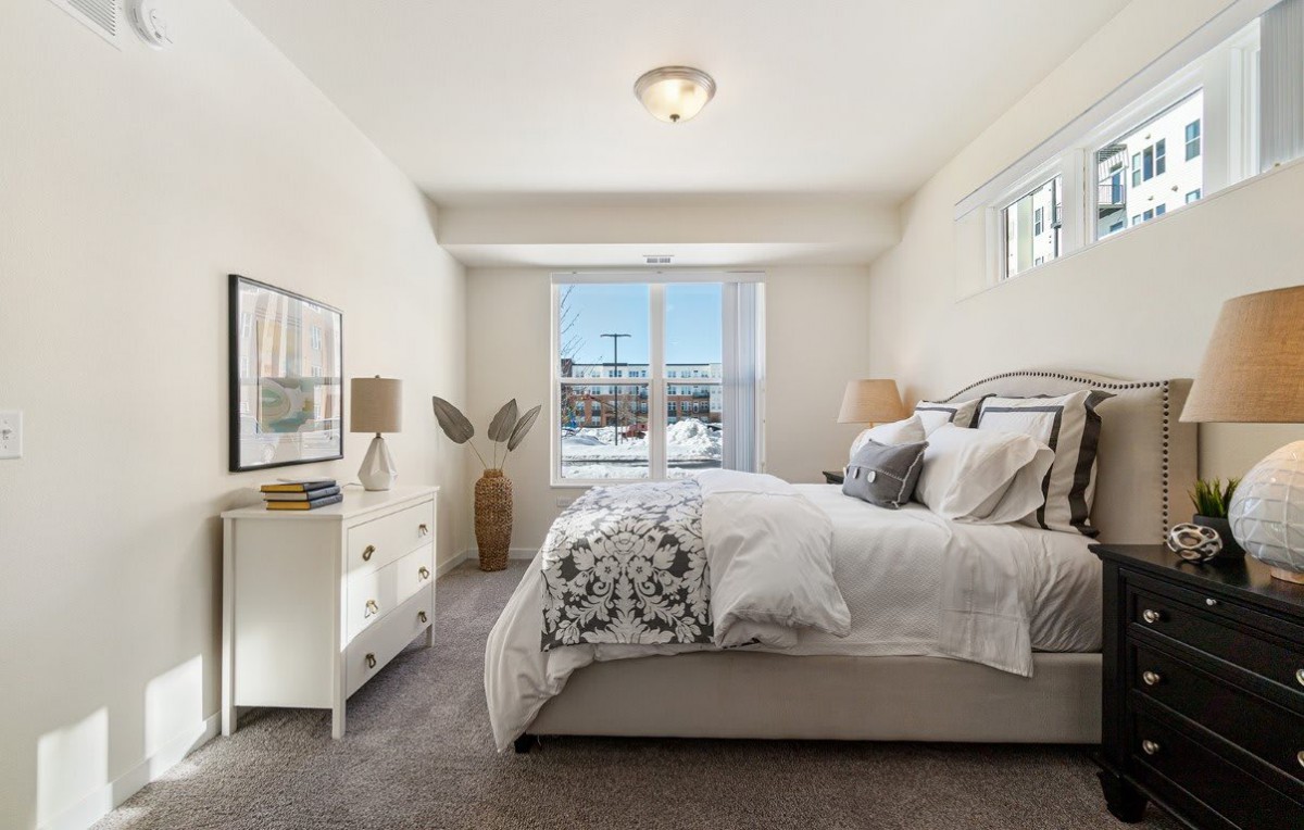 A cozy and stylish bedroom with a large window, featuring a comfortable bed, decorative pillows, two lamps on nightstands, and soft carpeting.