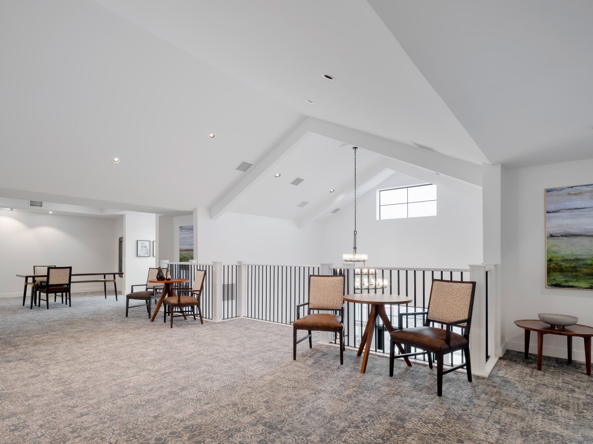 A spacious and bright loft area with seating and a table, featuring carpeted flooring and white railings.