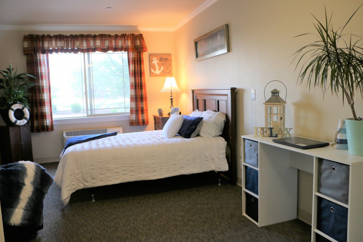 A cozy bedroom featuring a neatly made bed with pillows, a window with patterned curtains, and various decor items including a lamp and a small shelf.