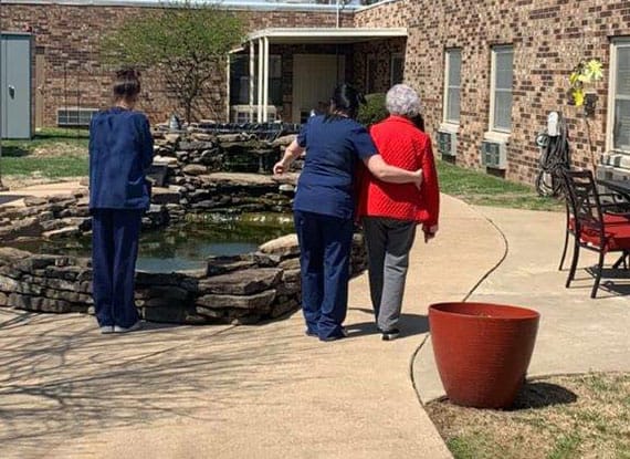 A healthcare worker supports an elderly person while walking outdoors near a building.