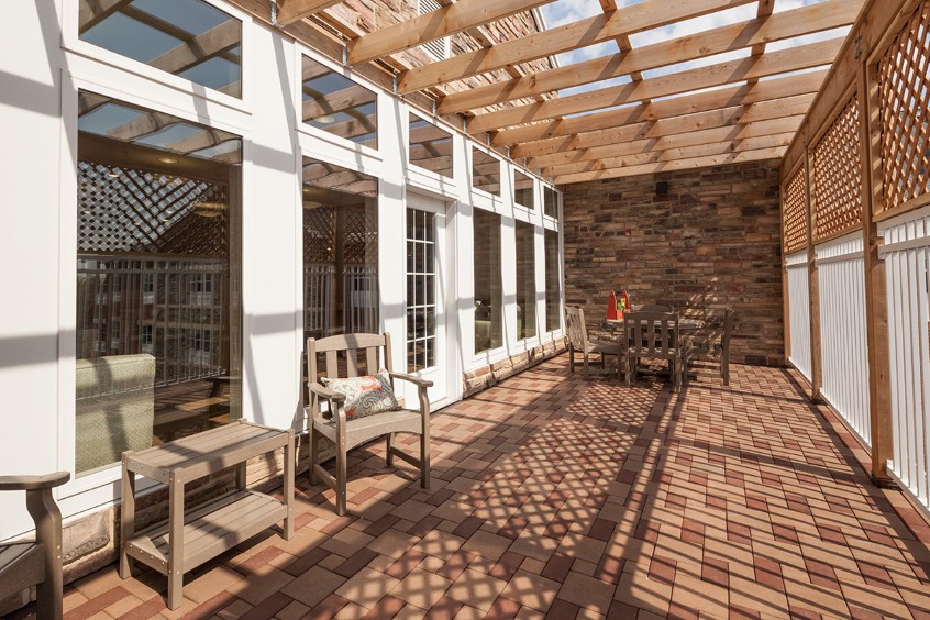 A sunlit patio with wooden furniture and a pergola overhead, featuring a brick floor.
