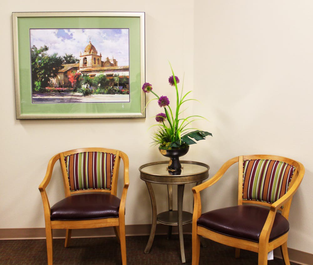 A waiting room with two chairs and a small table with a decorative plant, under a framed painting on the wall.