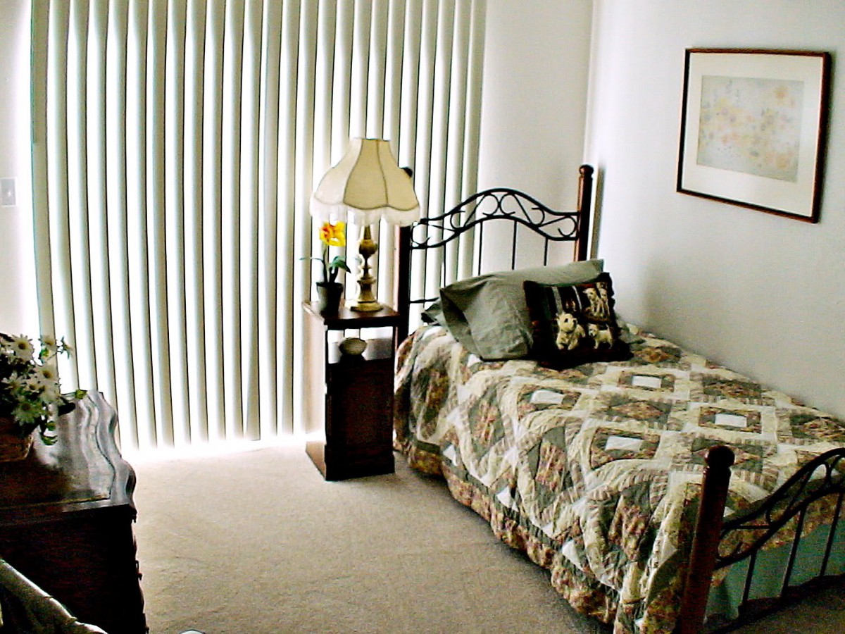 A cozy bedroom with a single bed, adorned with a patterned quilt, next to a bedside table and lamp. Vertical blinds cover the window area.