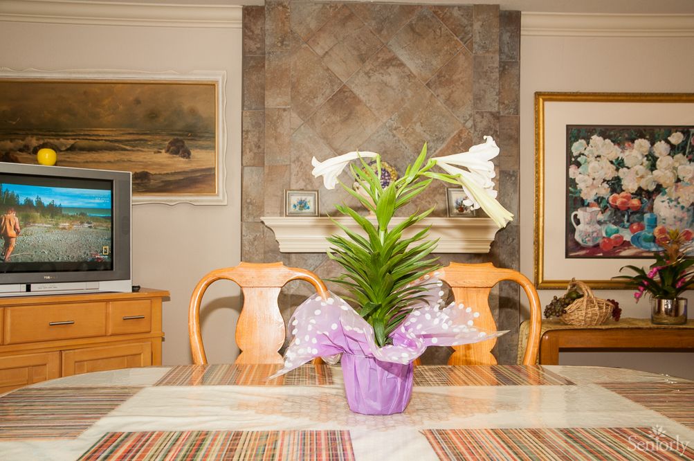 A dining room with a lily plant centerpiece on the table, surrounded by wooden chairs and decorated with artwork.