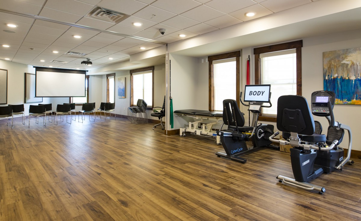 A spacious gym with wooden flooring and various exercise equipment near large windows.