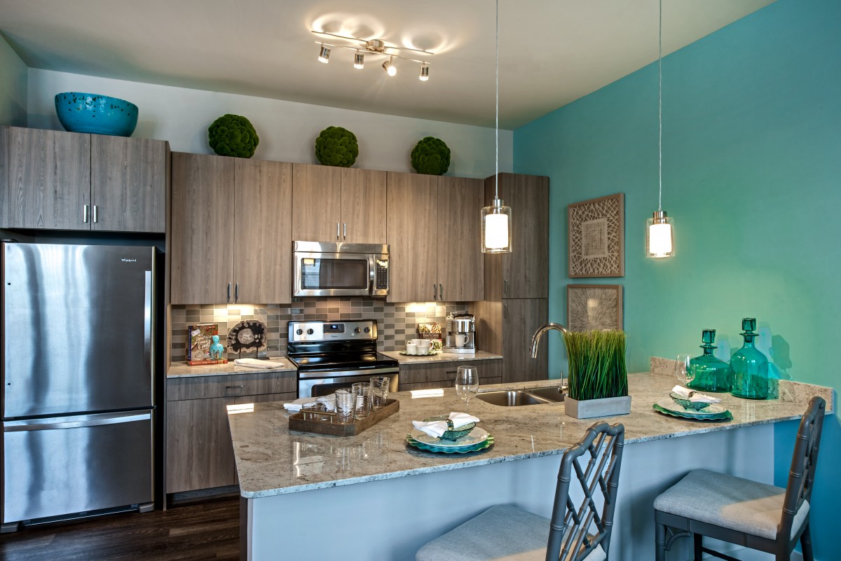 A modern kitchen featuring wood cabinets, granite countertops, and stainless steel appliances, accented by pendant lighting and decorative plants.