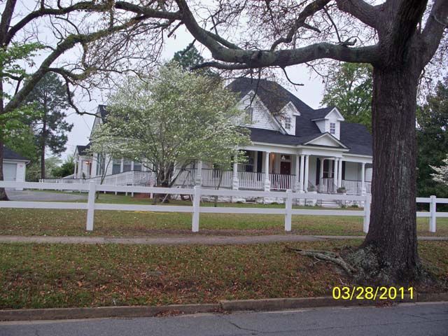 A picturesque house with a wrap-around porch, surrounded by trees and a white fence.
