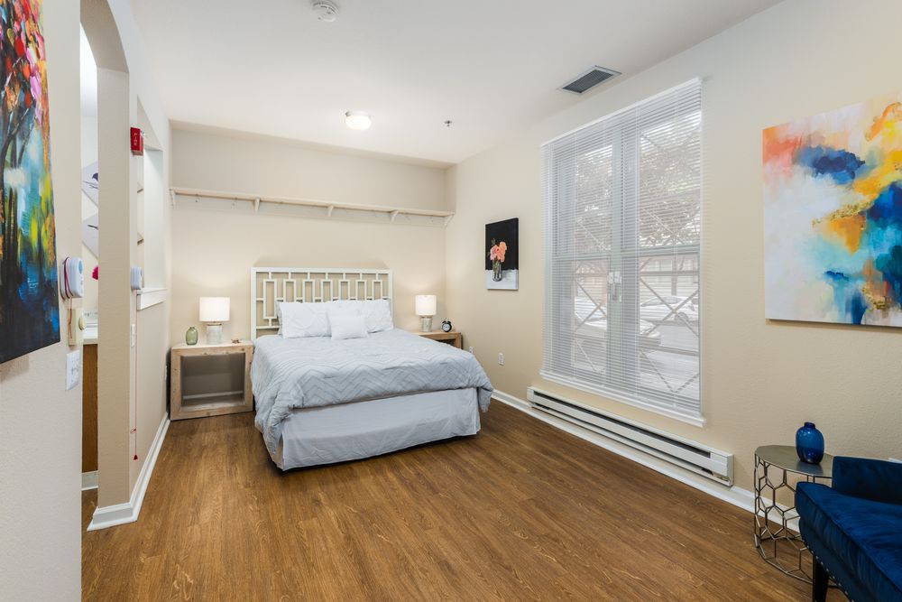 A cozy bedroom with a neatly made bed, wooden flooring, and large windows letting in natural light.