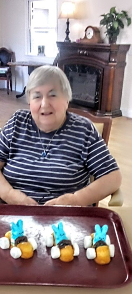 An elderly woman smiling and sitting indoors, wearing a striped shirt.