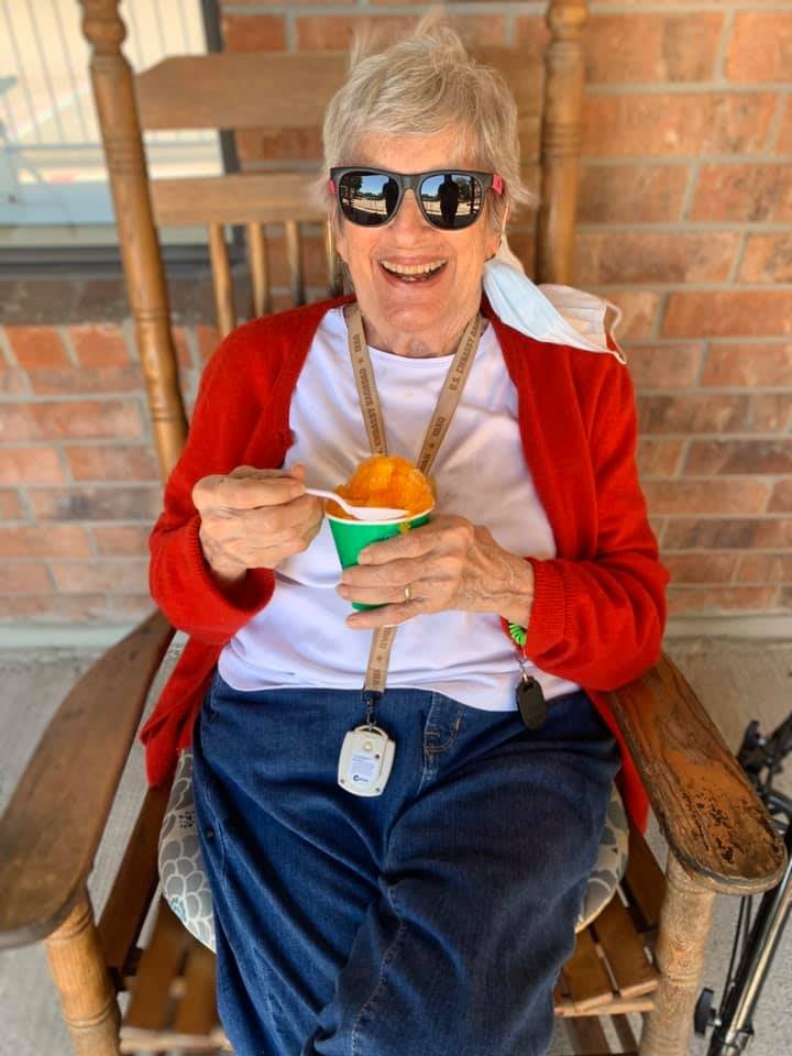 An elderly person wearing sunglasses and a red cardigan, enjoying a cup of ice cream while sitting on a rocking chair outdoors.