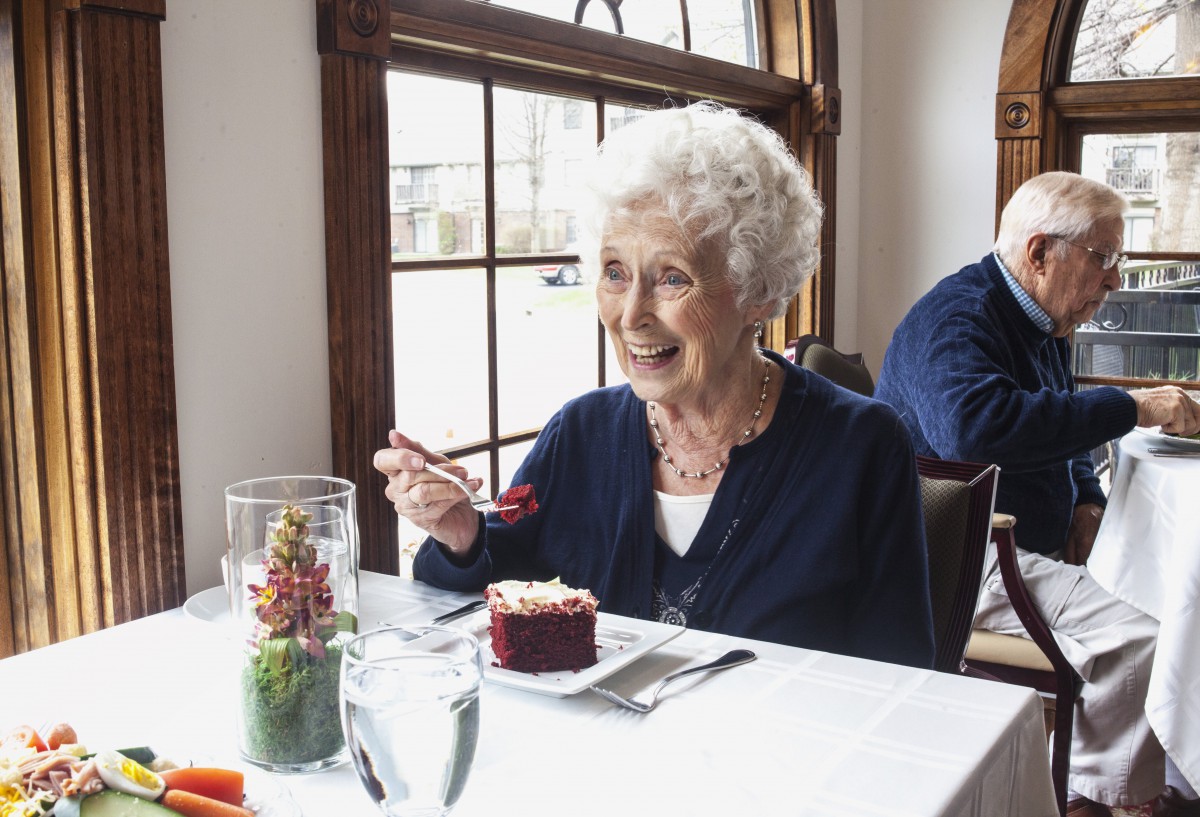 An elderly woman enjoying cake at a restaurant table by the window.