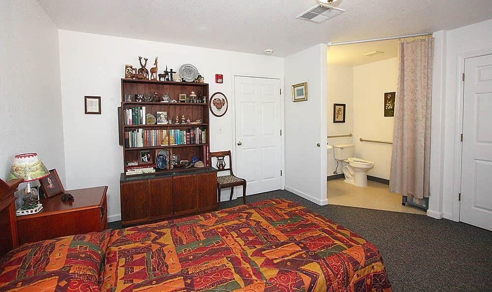A cozy bedroom featuring a colorful bedspread, a wooden bookshelf filled with books and decorations, and an adjoining bathroom.