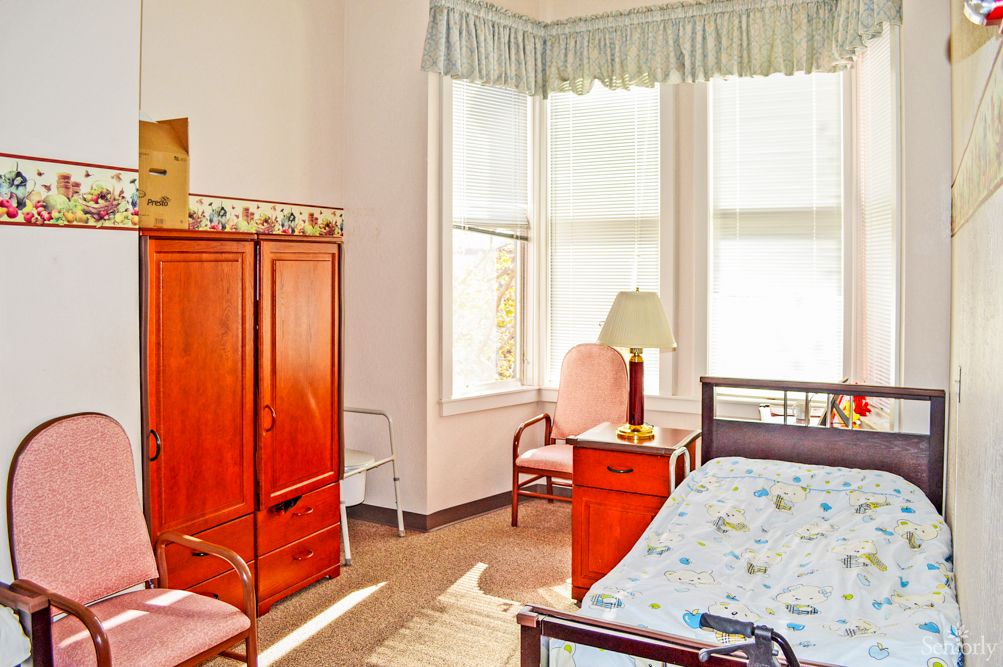 A bright and cozy bedroom with wooden furniture, including a bed, wardrobe, and chairs, illuminated by natural light from large windows.