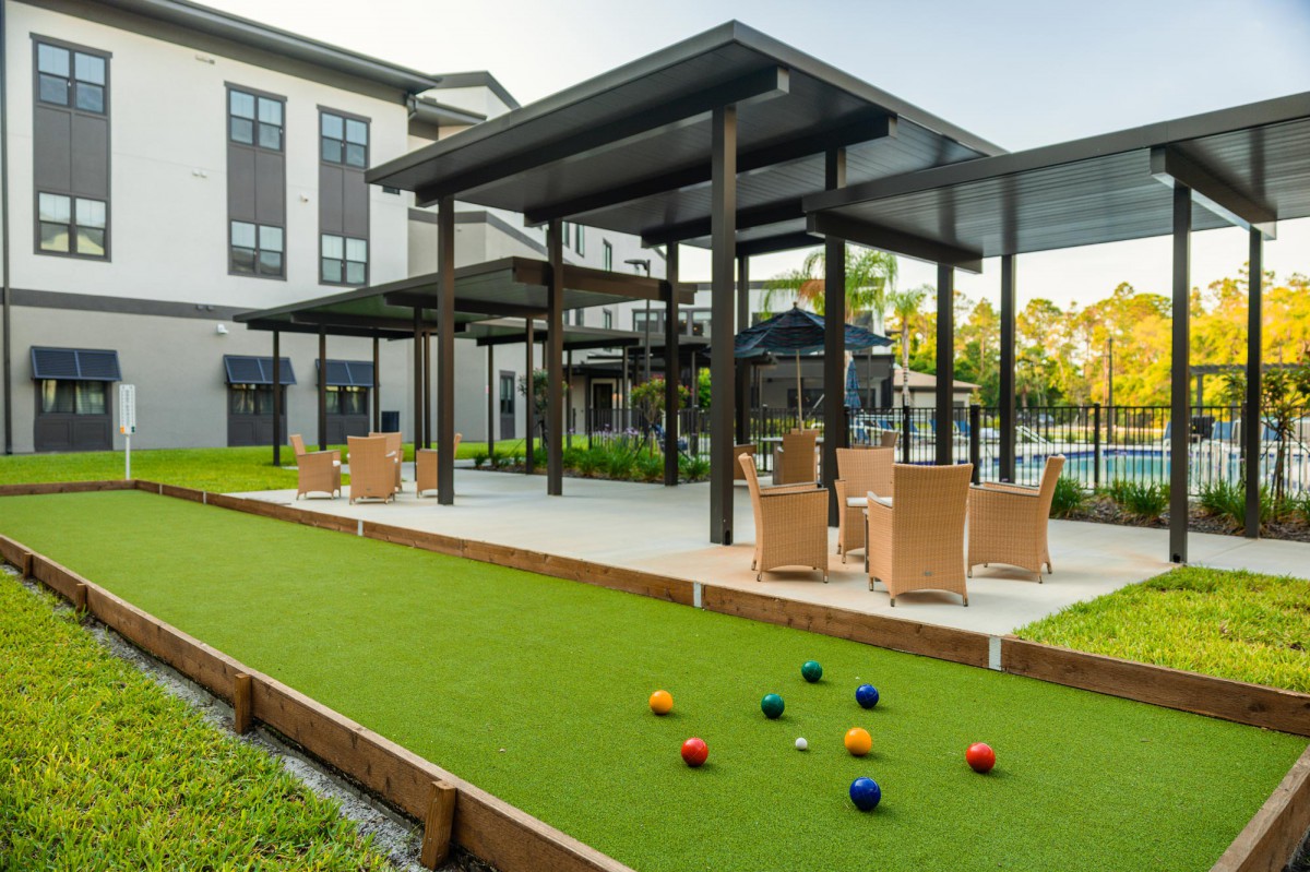 A modern outdoor patio with a pergola, seating area, bocce court, and landscaped surroundings next to a building.