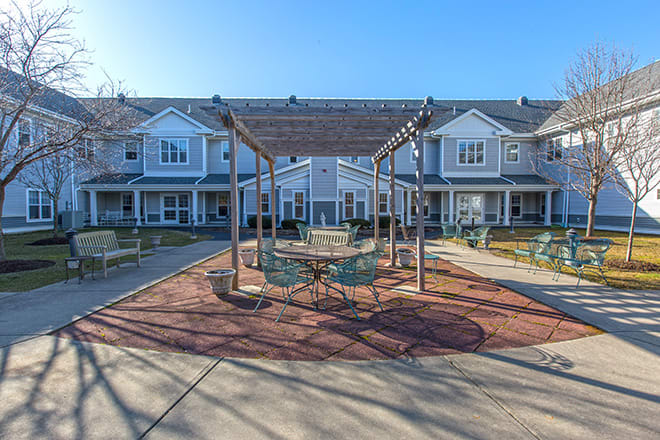 A sunny outdoor patio area with seating under a pergola in front of residential buildings.