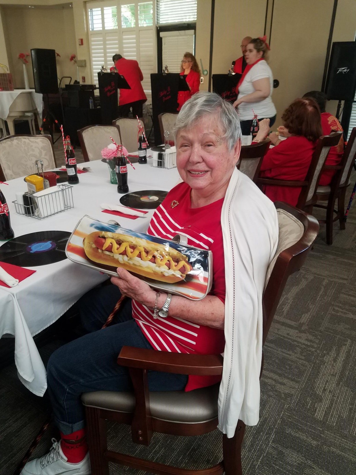 An elderly woman smiling while holding a decorated hot dog at a party.