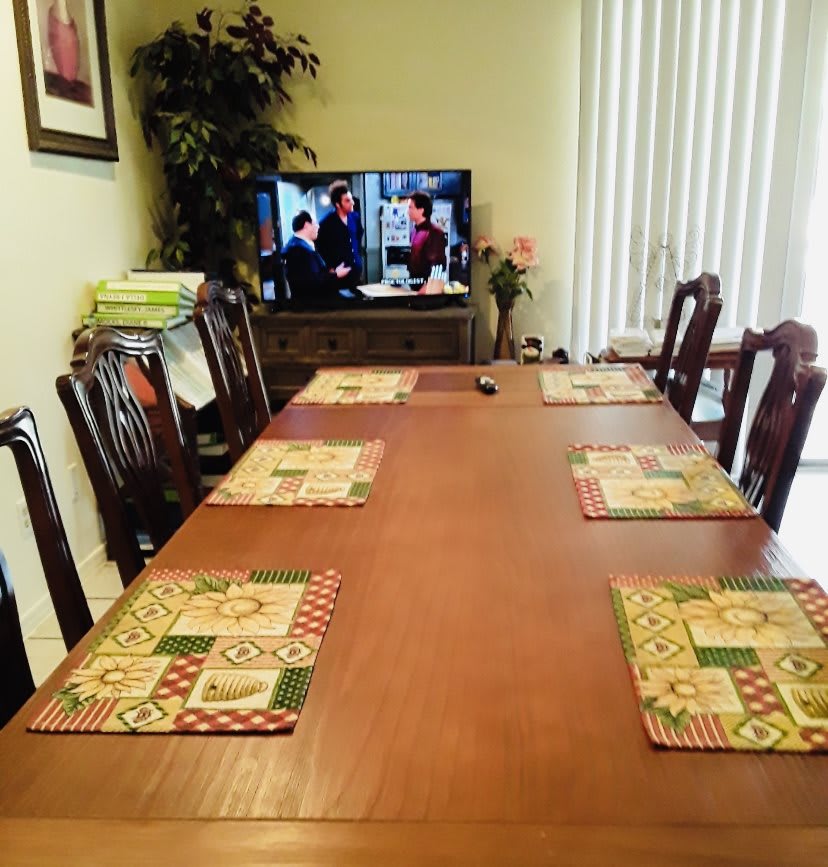 A dining room with a large wooden table, set with placemats and surrounded by chairs, facing a television.