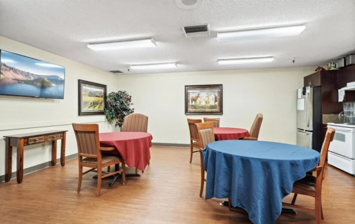 A well-lit dining room with tables covered in red and blue tablecloths, surrounded by wooden chairs and decorated with framed artwork on the walls.