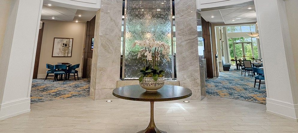 A stylish hotel lobby showcasing a round table with an elegant floral arrangement, set against a backdrop of marble walls and decorative glass.