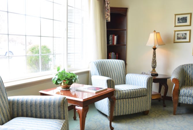 A cozy living room with striped armchairs, a wooden table, and a lamp near a large window.