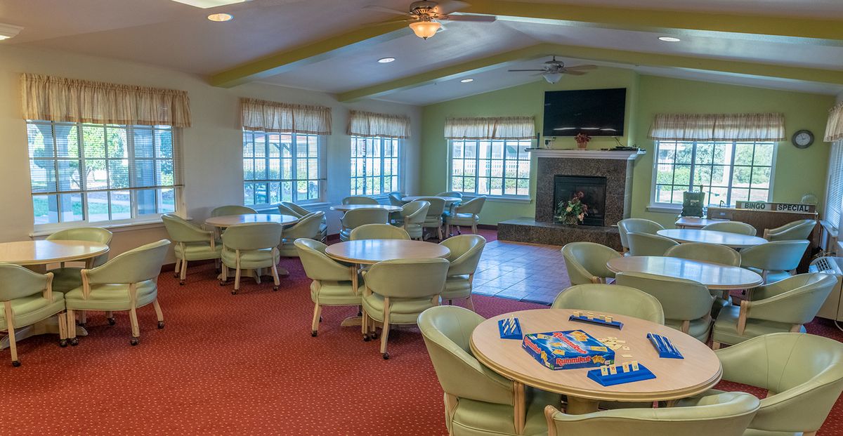 A spacious community room with tables and chairs, featuring a fireplace and large windows.